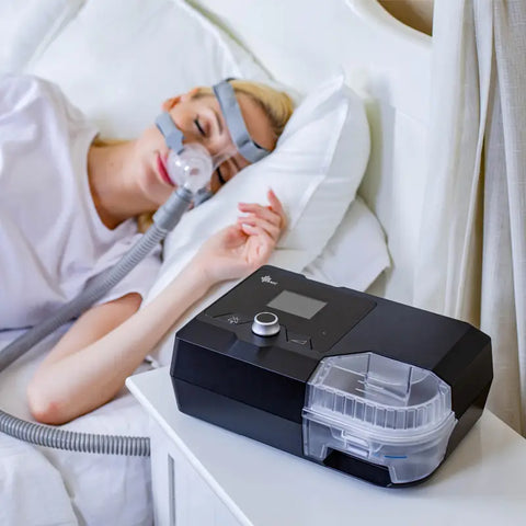  a woman using a sleep therapy machine, specifically designed to help with conditions like sleep apnea. She is in bed, lying down comfortably with a CPAP (Continuous Positive Airway Pressure) mask fitted over her nose and mouth, which is connected by a tube to the machine placed beside her. The sleep apnea device shown has a sleek black design with a clear water compartment and a digital display, visible on the bedside table.