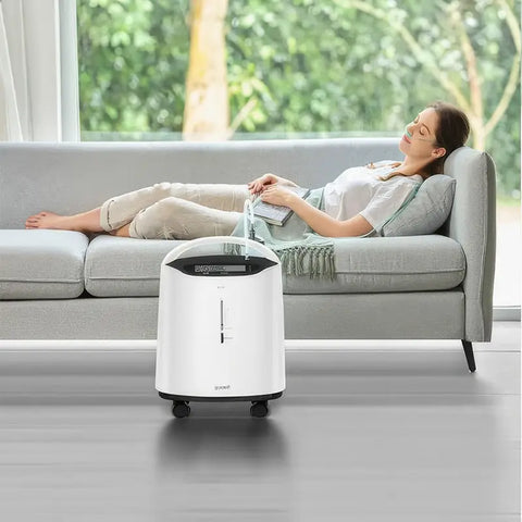  The device is primarily white with a sleek, modern design, featuring a handle on top for easy portability. It has a digital display at the top front, showing various readings and settings. Below the display, there is a flow rate indicator that ranges from 0.5 to 5 liters per minute. The oxygen concentrator is equipped with wheels, making it easy to move around. 
